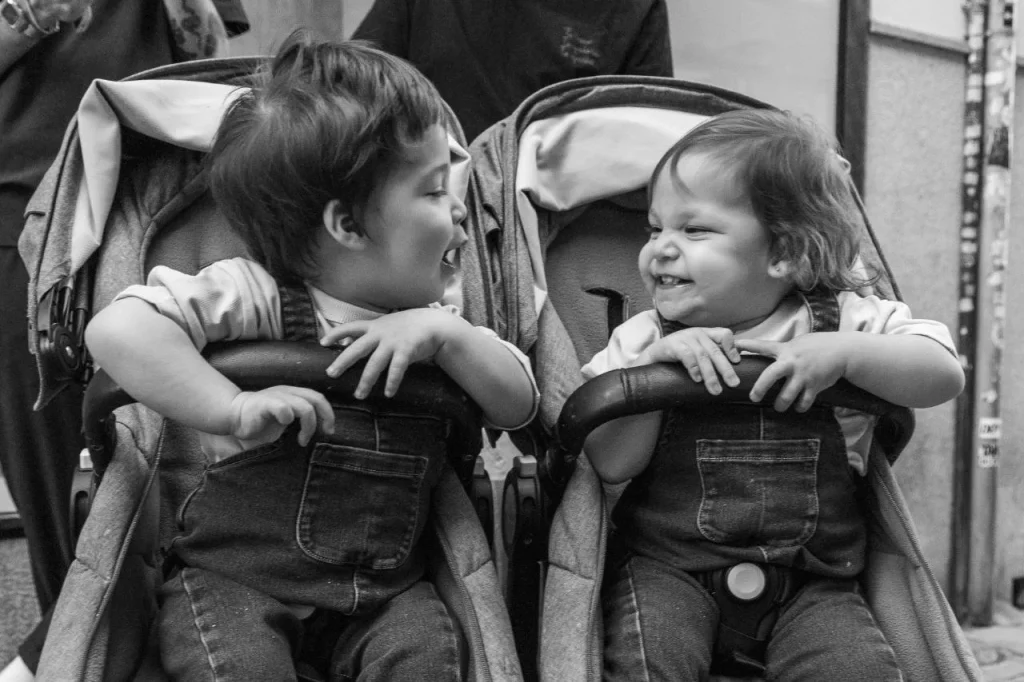 Two toddlers smiling at each other in a side-by-side double pushchair